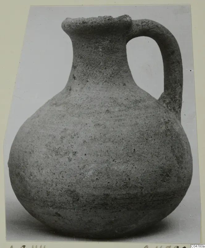 Pottery jug with a round body, narrow neck, flared rim and single strap handle, photographed against a pale background.
