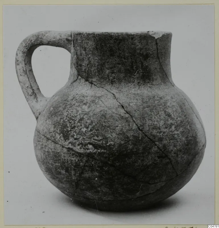 Round-bodied jug with a single loop handle and a short flared neck, cracked, photographed against a plain background.
