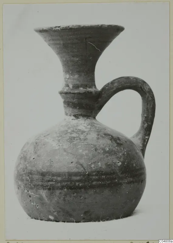 Vessel with a flaring neck, single loop handle and dark horizontal bands on the body, shown alone against a pale backdrop