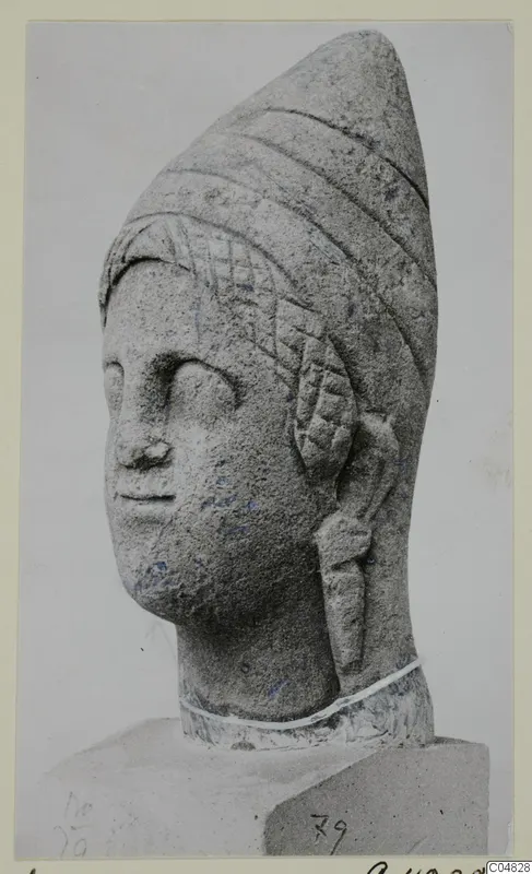 Stone statue head from Arsos with a tall cone-shaped headdress, shown in three-quarter view on a block base.