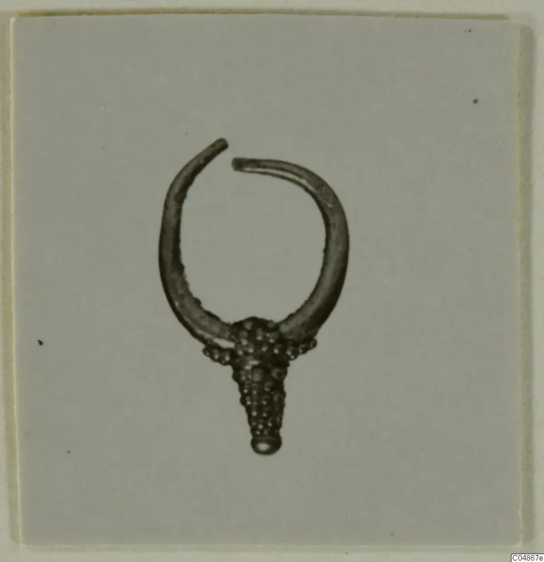 Earring of gold or silver-coloured metal, open ring with a granulated cone-shaped pendant, photographed on a plain grey card.