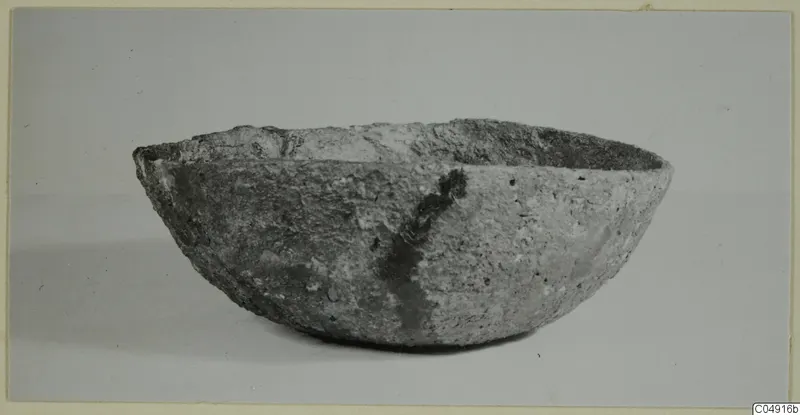 Side view of a thick-walled, rounded bowl with an uneven rim and a pitted, pale grey surface