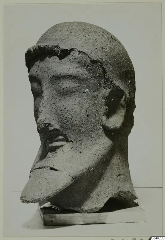 Sculpted head with a moustache and broken chin, from Mersinaki, shown on a wooden block