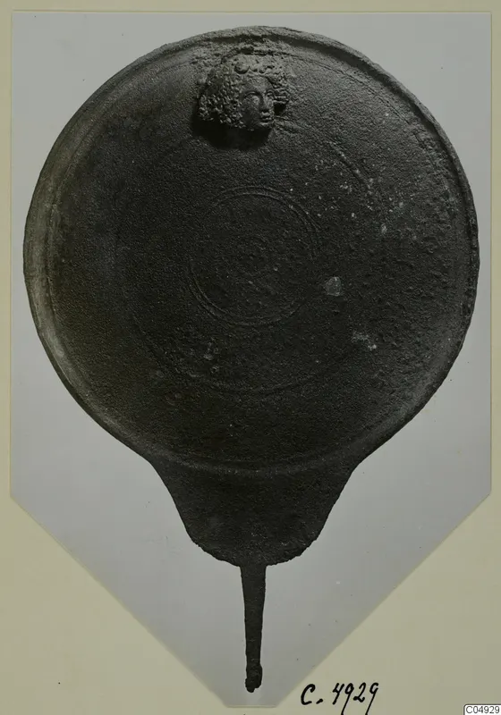 Round dark metal disc with a long tang and a small relief head of a figure with curly hair, from Marion