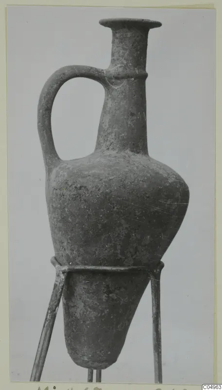 Base-ring ware juglet with a long neck, flared rim and loop handle, shown on a metal stand.