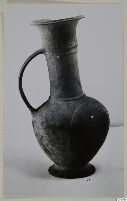 Pottery Base-Ring Ware jug with a single strap handle and flaring rim, shown standing on a pale surface