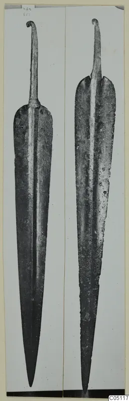 Two long leaf-shaped metal blades with curled tangs, photographed upright side by side.