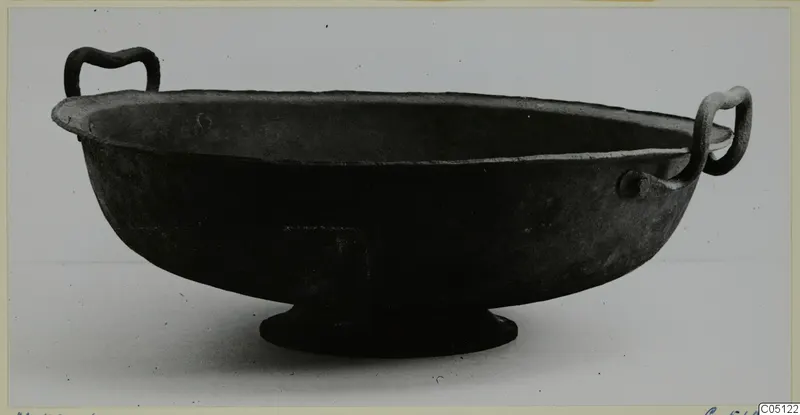 Dark metal bowl with two handles and a low ring foot, photographed on a pale background