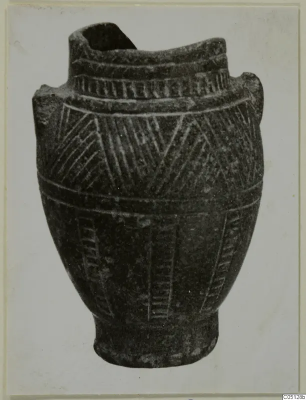 Dark stone vessel with incised hatched decoration and a broken rim, photographed on a light card