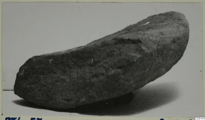 Stone tool from Petra tou Limniti, dark grey and elongated, photographed on a pale background