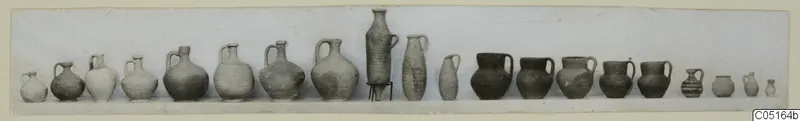 Row of about twenty pottery jugs and jars from Kountoura Trachonia, Tomb 8, set side by side on a pale surface