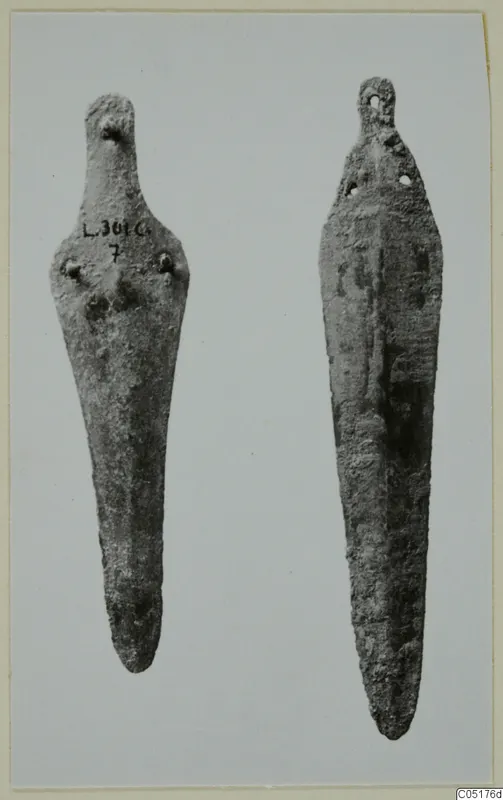 Two corroded bronze knives side by side against a pale background, one with a handwritten number label