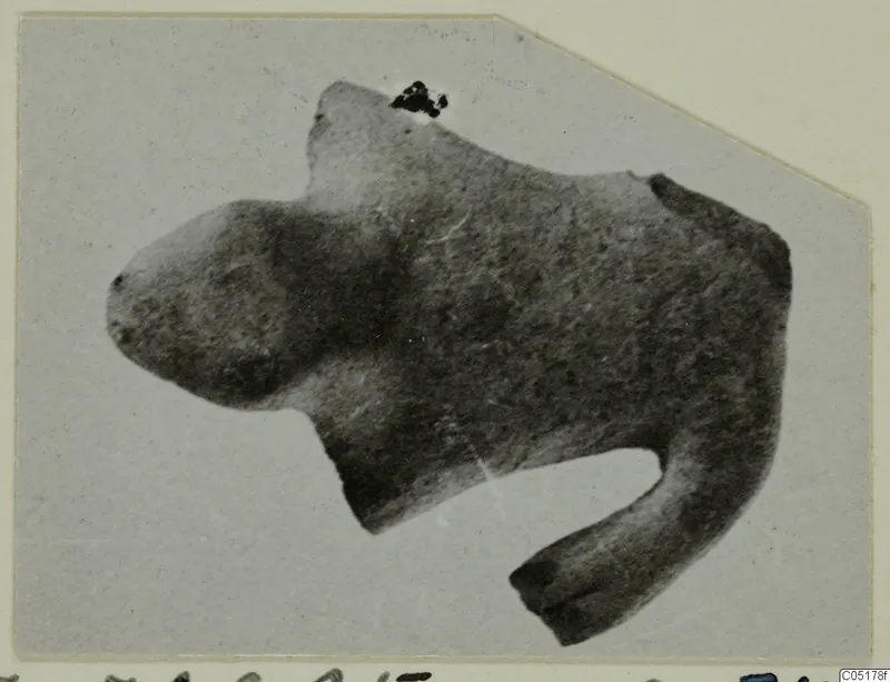 Terracotta figurine of an animal with a blunt head and one leg extended downward, shown as a mounted print.