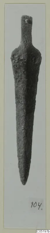 Corroded metal tool with a narrow tang at the top and a long tapering point, shown on a photo mount.