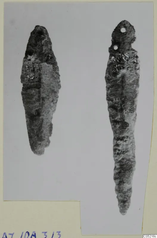 Two corroded metal tools or weapons from Ayios Iakovos Tomb 10 A, one with two small holes near its top end.