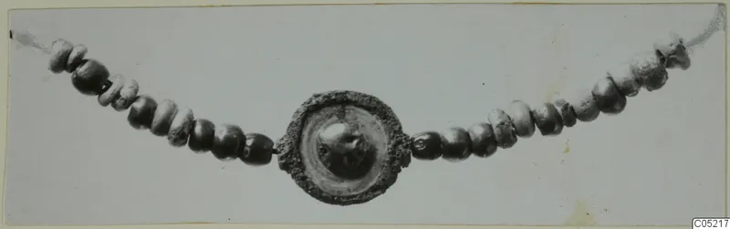 Necklace of small beads strung on a cord, with a round disc pendant with a raised central boss at the centre, on a pale card.