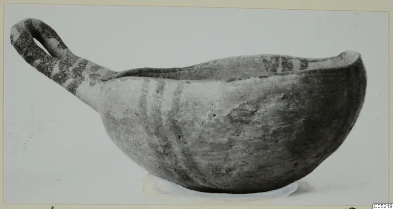 Pottery bowl with a long strap handle and dark painted bands, photographed against a plain background.