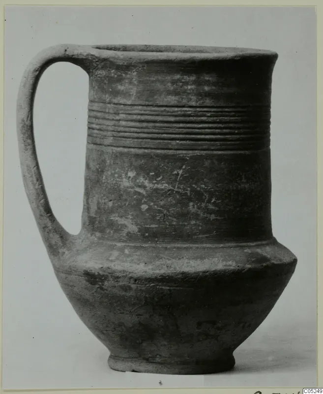 Handled pottery vessel with a tall body, a band of horizontal grooves and a flared rim, dark grey-brown surface