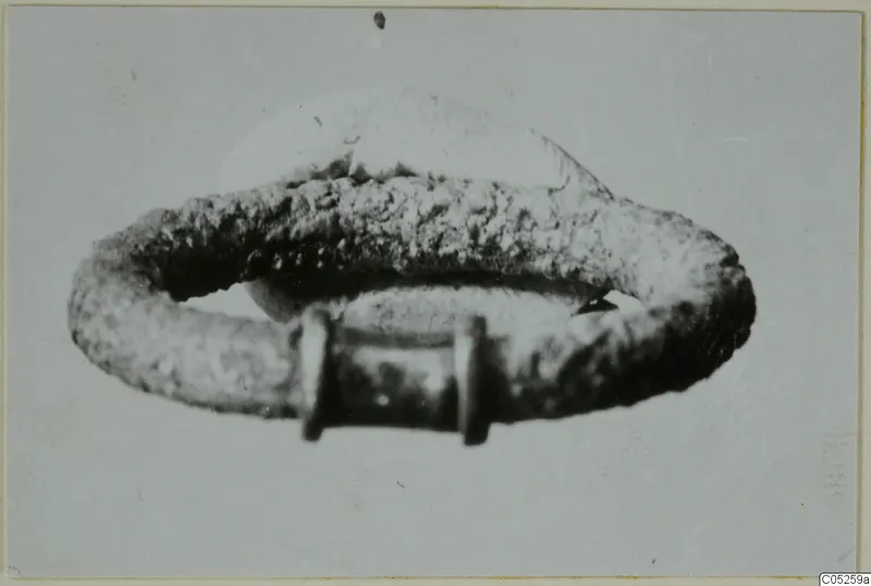 Ring of corroded metal with a flat raised oval plate on top and a rectangular fitting at the front
