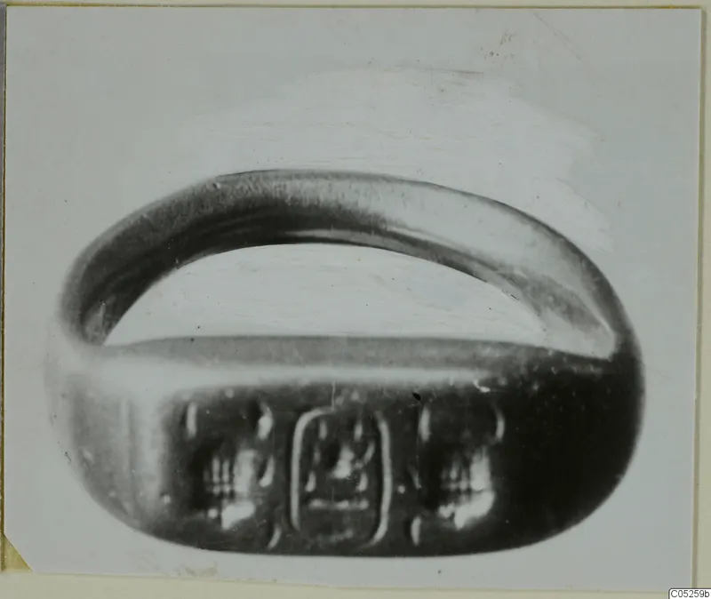 Metal finger ring with a wide oval bezel bearing an engraved design, shown on a pale photo mount.