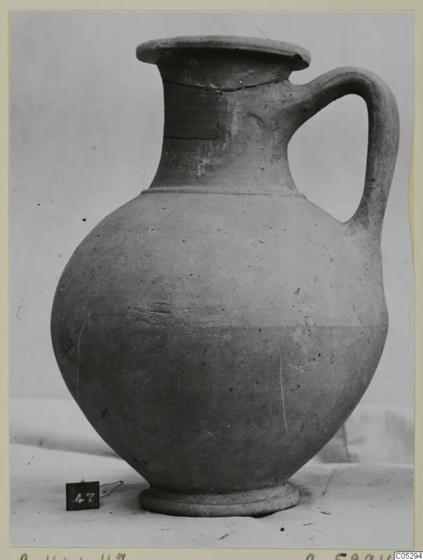 Plain pottery jug with a single handle and a flared rim, shown on a white cloth against a pale backdrop.