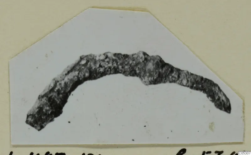 Corroded, curved bronze object from Lapithos Tomb 403, shown on a pale mounted card