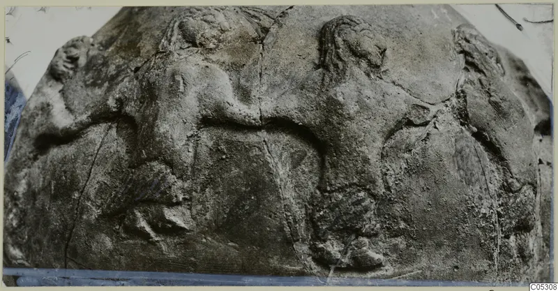 Close view of a weathered stone surface with raised relief figures, including two heads and arms, cracked across the middle