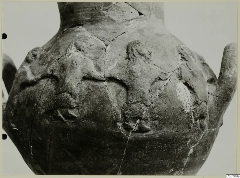 Close view of the body of a large pottery vessel with applied relief figures and cracks across the surface