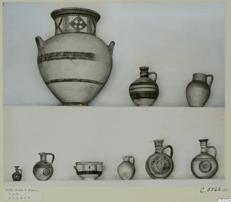 Pottery vessels from Stylli, Tomb 8, arranged on two white shelves against a plain background
