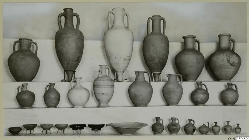 Pottery from Marion, Tomb 22: tall amphorae, jugs and small bowls arranged in rows on a light backdrop.