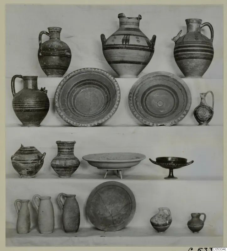 Many pottery vessels, jugs, jars, dishes and a two-handled cup, arranged in four rows on white-draped shelves.