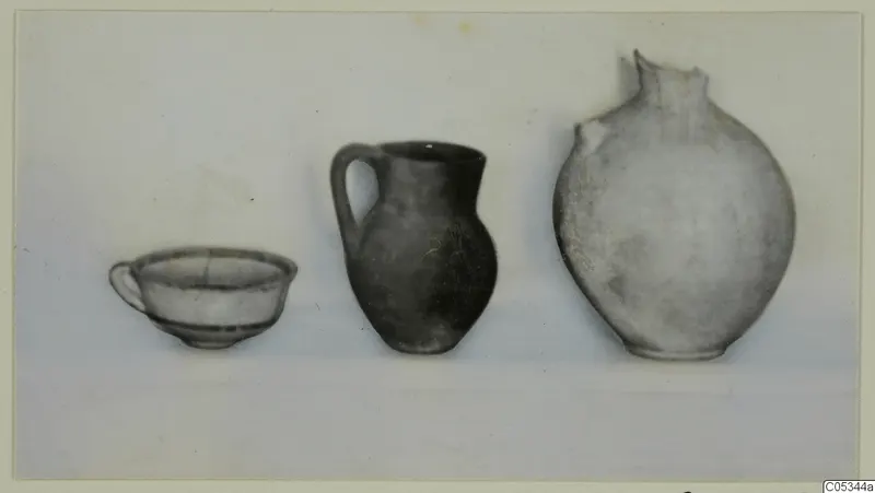 Three pottery vessels in a row: a small bowl with a handle, a dark jug with a strap handle, and a large rounded vessel with a broken rim.