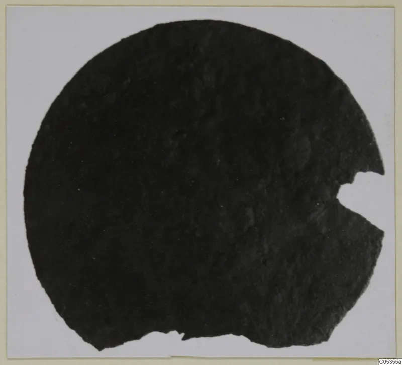 Dark, corroded circular object with a broken notch on its right edge, shown on a pale card. Marion, Tomb 72.