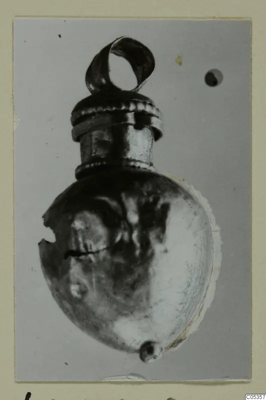 Small metal object with a loop at the top and a ribbed collar, photographed on a card mount.