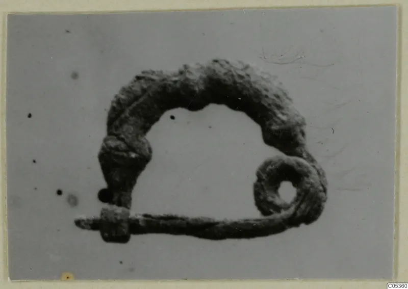 Bronze fibula from Amathus with a coiled spring and long pin, photographed against a plain grey background.