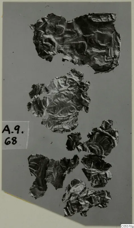 Crumpled, thin metal fragments scattered on a grey mount, with a handwritten number label at the left edge