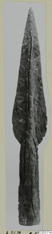 Bronze spearhead with a long leaf-shaped blade and a central rib, photographed upright against a grey backdrop.