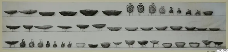 Three rows of pottery bowls, dishes and small jugs set out on shelves against a pale background.