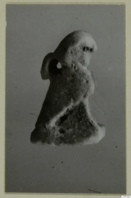 Small figure with a suspension hole through a lug at its side, shown against a grey backdrop