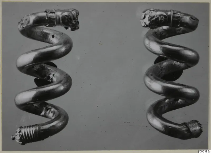 Two spiral gold objects, each with a moulded head-like terminal at the top, photographed side by side on a grey ground.