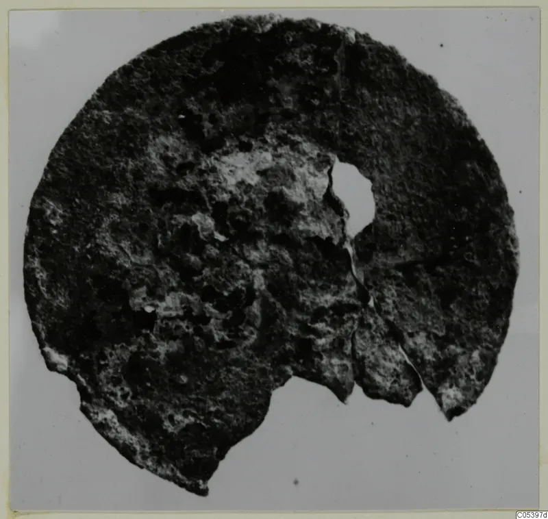 Corroded bronze disc fragment with an off-centre hole, from Marion Tomb 9, shown on a grey background.
