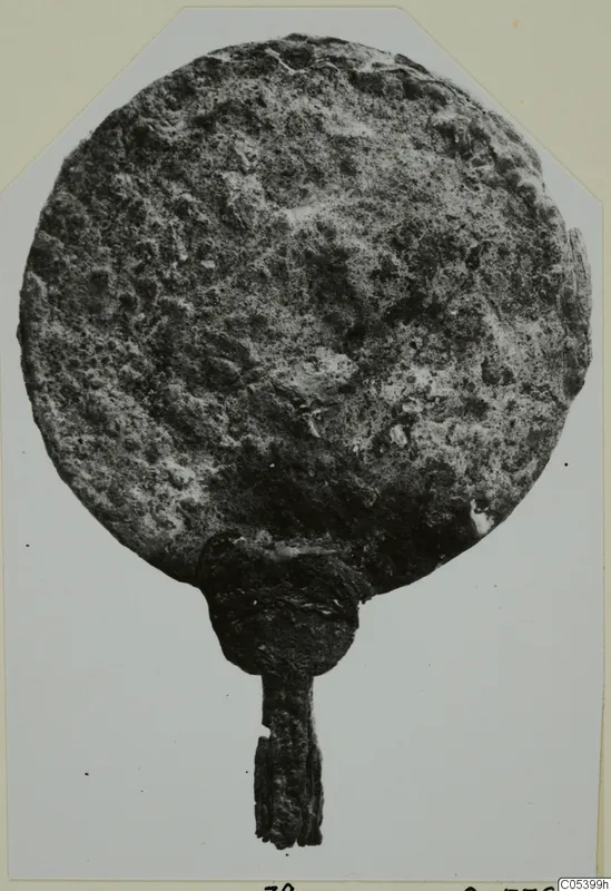 Corroded bronze disc with a short handle, from Marion tombs 61–98, photographed face-on on a mounted card.