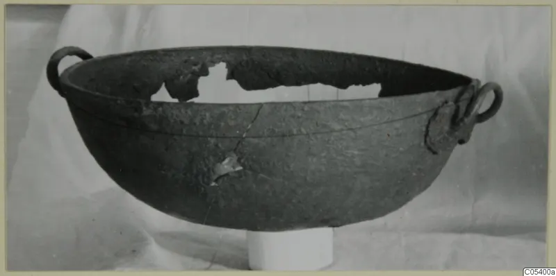 Dark, corroded metal bowl with two loop handles, shown on a white pedestal