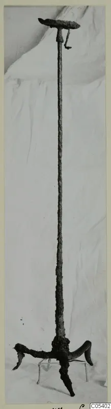 Corroded iron object with a flat disc top, long shaft and three curved feet, standing upright on white cloth