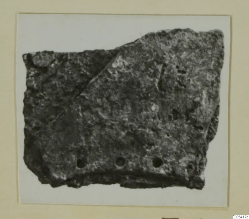 Fragment of a dark, encrusted object with three round holes along its lower edge, from Amathus tombs 2-26.