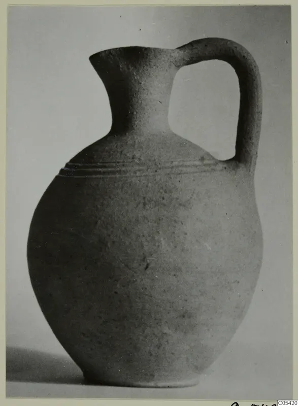 Plain pottery jug with a rounded body, a flared lip and a single strap handle, photographed on a grey surface.