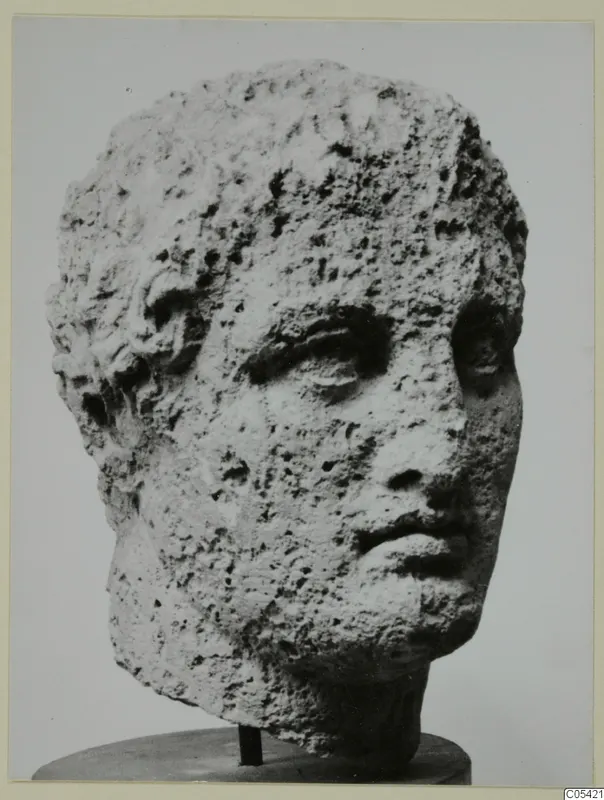 Stone head of a figure from Mersinaki, with short hair and a heavily pitted surface, photographed on a round stand.