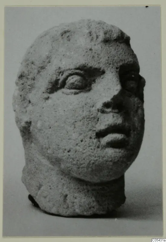 Stone head from Mersinaki with large eyes, a broken nose and a pitted surface, seen against a grey backdrop