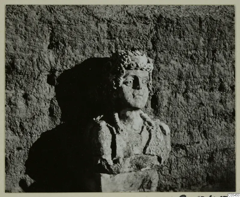 Carved stone bust of a figure with a wreath or curled headdress, set against a rough textured wall.