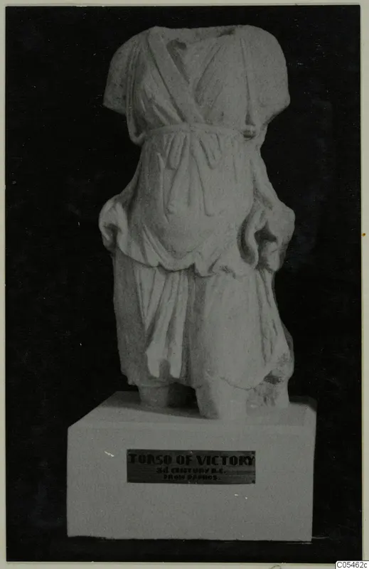 Marble sculpture of a draped standing figure, headless and armless, on a white plinth against a black background.
